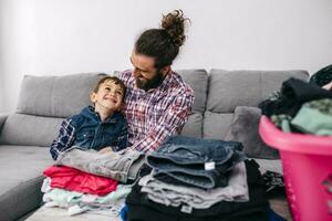 padre e figlio seduta insieme su il divano avendo divertimento mentre pieghevole lavanderia foto