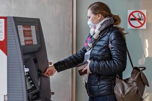 una donna che indossa una maschera medica protettiva sul viso che inserisce una carta di credito e preleva contanti da un bancomat. foto