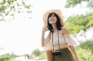 ritratto di asiatico giovane donna viaggiatore con tessitura cappello e cestino e un' telecamera su verde pubblico parco natura sfondo. viaggio viaggio stile di vita, mondo viaggio esploratore o Asia estate turismo concetto. foto