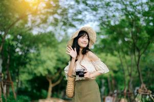 ritratto di asiatico giovane donna viaggiatore con tessitura cappello e cestino e un' telecamera su verde pubblico parco natura sfondo. viaggio viaggio stile di vita, mondo viaggio esploratore o Asia estate turismo concetto. foto