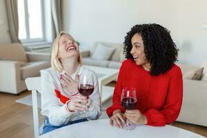 donne seduta su divano ridendo nel un' accogliente soffitta appartamento con vino. Due femmina amici rilassante su divano a casa con bicchiere di vino parlando insieme. foto