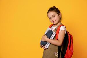 caucasico adorabile inteligente primario alunno ragazza trasporto un arancia zaino, Tenere scuola forniture, sorridente a telecamera foto