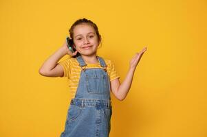 carino poco ragazza parla su mobile Telefono, sorrisi con bellissimo trentadue denti Sorridi guardare a telecamera, isolato giallo sfondo foto