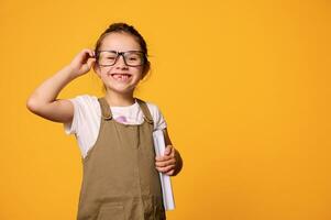 caucasico malizioso bambino ragazza, primo grado alunno Tenere libro e sorridente guardare a telecamera attraverso sua occhiali foto