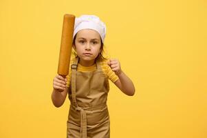 grave arrabbiato poco bambino ragazza nel dello chef uniforme, Tenere un' di legno rotolamento spillo, stringendo pugni, guardare a telecamera foto