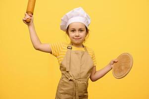 bellissimo bambino ragazza nel capocuoco cappello e grembiule, sorridente a telecamera, Tenere rotolamento perno e di legno asse, isolato su giallo foto