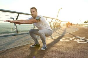 di mezza età atleta, sportivo è impegnato nel gli sport su un' città ponte a alba, esegue in profondità squat, impasta muscoli di il corpo. concetto di sport, movimento, energia e dinamico, salutare stile di vita. foto