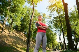 parte inferiore Visualizza di un' fiducioso 30 anni vecchio bella donna nel abbigliamento sportivo in piedi nel un' foresta città parco con sua braccia attraversato e sorridente con un' bellissimo trentadue denti Sorridi durante sua mattina correre o allenarsi foto