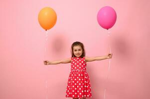 adorabile bellissima 4 anni bambino ragazza nel rosa vestito con bianca polka punti, Tenere colorato palloncini nel sua disteso mani, sorridente guardare a il telecamera, isolato su rosa sfondo con copia spazio foto