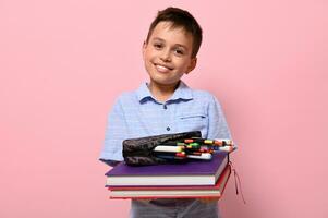 un' sorridente alunno a scuola, detiene libri nel davanti di lui e matita Astuccio con penne, pennarello penne e marcatori caduta su di lui. indietro per scuola concetti su rosa sfondo con copia spazio foto