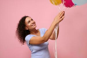 studio tiro per anno Domini di allegro bella donna raggiungendo per multicolore aria palloncini sorridente felicemente contro rosa sfondo con copia spazio. il concetto di celebrazione, vacanze, anniversario e i regali. foto