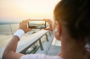 posteriore Visualizza di giovane atleta donna nel rosa maglietta Tenere smartphone e assunzione foto di bellissimo Alba a il fine di il ponte prima mattina fare jogging. mobile Telefono nel vivere Visualizza modalità. all'aperto allenarsi