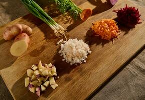 triturati barbabietola, grattugiato carota, cipolla e cubi di tagliare Patata su un' di legno tavola con metà di verdure con verde le foglie e radici su il sfondo. avvicinamento foto