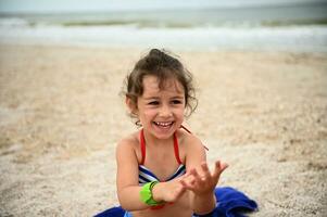 fiducioso ritratto di bellissimo bambino ragazza riposo su il spiaggia, carino sorridente in posa per telecamera contro sabbioso spiaggia sfondo. estate vacanze concetto foto