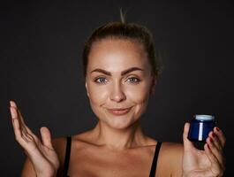 bellissimo giovane bene guardare donna con fresco raggiante chiaro pelle sorrisi guardare a telecamera e Tenere un' bottiglia con anti età idratante facciale crema, isolato al di sopra di nero sfondo con copia spazio foto