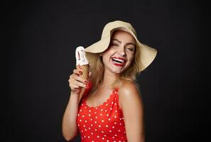 attraente sexy allegro sbalorditivo bionda donna nel un' costume da bagno e estate cannuccia cappello gioisce, Tenere cornetta ghiaccio crema nel sua mani, sorridente allegramente con bellissimo trentadue denti Sorridi al di sopra di nero sfondo foto