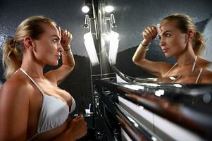 attraente europeo giovane donna massaggio sua viso con pietra massaggiatore, guardare a sua riflessione nel il bagno specchio foto
