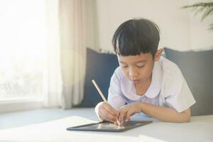 contento asiatico ragazzo 6-7 anni vecchio, nero capelli, bianca pelle, indossare bianca camicia e blu pantaloncini scrittura su un' lavagna. bambini nel scuola uniformi di Tailandia. concetto di formazione scolastica, giocare a, creatività. foto