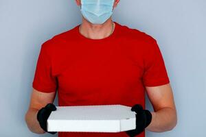 consegna uomo dipendente nel rosso berretto vuoto maglietta uniforme viso maschera guanti hold cartone scatola Pizza su grigio sfondo servizio quarantena pandemia coronavirus virus 2019-ncov concetto. foto