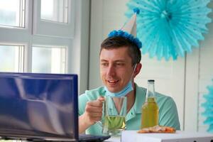 un' uomo nel un' medico maschera si siede nel davanti di un' il computer portatile e onde ciao. in linea compleanno. foto