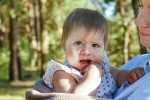 bambino nel sua La madre di braccia detiene sua dito nel sua bocca nel il parco su un' soleggiato giorno foto