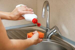 donna di mani versare detergente su un' spugna al di sopra di un' Lavello con un' acqua rubinetto foto