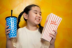 asiatico bambino ragazza mangiare Popcorn e potabile bibita su giallo parete sfondo, cinema concetto, foto