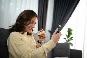 asiatico donna utilizzando Telefono video chiamata con amico mentre mangiare Sandwich a casa foto