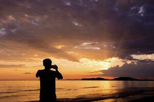 un' uomo in piedi su il spiaggia assunzione un' foto di il tramonto