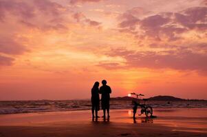 silhouette coppia in piedi su il spiaggia con un' bicicletta a tramonto foto