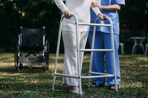 asiatico giovane donna infermiera a assistenza infermieristica casa prendere cura Disabilitato anziano uomo. badante medico servire fisico terapia per più vecchio anziano paziente per esercizio e pratica a piedi su camminatore o canna a Giardino dietro la casa. foto