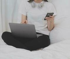 efficiente giovane del Millennio ragazza seduta su un' letto nel il mattina, usi il computer portatile computer e mangia Cornetti e bevande caffè per prima colazione foto