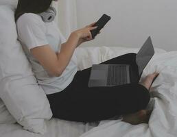 efficiente giovane del Millennio ragazza seduta su un' letto nel il mattina, usi il computer portatile computer e mangia Cornetti e bevande caffè per prima colazione foto