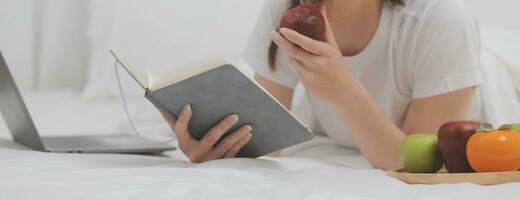 efficiente giovane del Millennio ragazza seduta su un' letto nel il mattina, usi il computer portatile computer e mangia Cornetti e bevande caffè per prima colazione foto