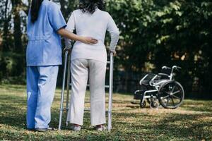 asiatico giovane donna infermiera a assistenza infermieristica casa prendere cura Disabilitato anziano uomo. badante medico servire fisico terapia per più vecchio anziano paziente per esercizio e pratica a piedi su camminatore o canna a Giardino dietro la casa. foto