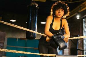 donna combattente ragazza potenza. africano americano donna combattente con boxe guanti in piedi su boxe squillare pendente su corde in attesa e riposo. forte potente ragazza. forza in forma corpo allenarsi addestramento. foto