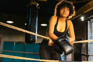 donna combattente ragazza potenza. africano americano donna combattente con boxe guanti in piedi su boxe squillare pendente su corde in attesa e riposo. forte potente ragazza. forza in forma corpo allenarsi addestramento. foto