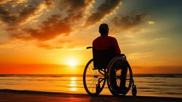 silhouette di Disabilitato uomo su sedia a rotelle a il spiaggia durante tramonto, disattivare giorno foto