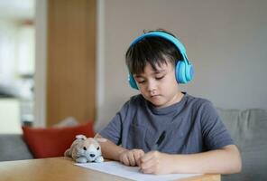 contento ragazzo indossare alcuni cuffie e ascoltando per musica mentre disegno su carta, interno ritratto di carino ragazzo godere un' creativo attività a casa su un' fine settimana. un' bambino è fare compiti a casa foto