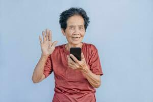 asiatico anziano donna sorrisi e video chiamata isolato nel grigio sfondo. forte anziano nonna sensazione contento utilizzando mobile cellula Telefono per comunicare con famiglia e godere la pensione vita nel Casa. foto