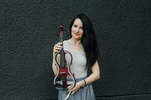 un' donna artista con buio capelli nel un' vestito detiene un' di legno concerto elettrico violino nel sua mani foto