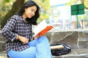 contento e succes femmina asiatico Università alunno godendo leggere un' libro nel il parco foto