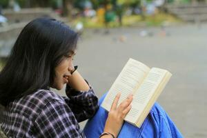 contento e succes femmina asiatico Università alunno godendo leggere un' libro nel il parco foto