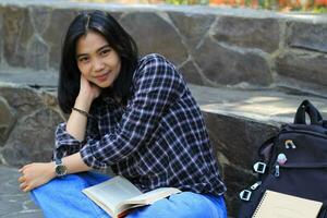 contento giovane asiatico donna Università alunno con sorridente viso godere leggere un' libro per esame nel il parco foto