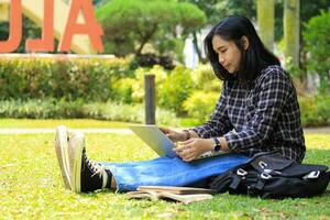 ritratto di focalizzata giovane asiatico donna fare surf Internet utilizzando il computer portatile per Lavorando a distanza, leggere prenotare, e scrittura nel taccuino. attraente asiatico alunno studia nel all'aperto foto