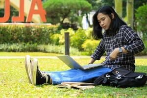 ritratto di focalizzata giovane asiatico donna fare surf Internet utilizzando il computer portatile per Lavorando a distanza, leggere prenotare, e scrittura nel taccuino. attraente asiatico alunno studia nel all'aperto foto