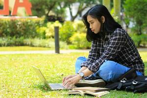 ritratto di focalizzata giovane asiatico donna fare surf Internet utilizzando il computer portatile per Lavorando a distanza, leggere prenotare, e scrittura nel taccuino. attraente asiatico alunno studia nel all'aperto foto