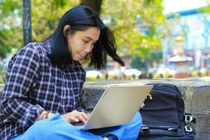 contento giovane asiatico donna focalizzata utilizzando il computer portatile Lavorando remoto e navigazione nel sociale media nel confortevole all'aperto spazio foto