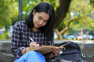sorridente asiatico bellissimo giovane donna godere scrittura per fare elenco e idea nel taccuino nel all'aperto città parco foto