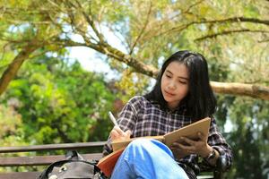 sorridente asiatico bellissimo giovane donna godere scrittura per fare elenco e idea nel taccuino nel all'aperto città parco foto